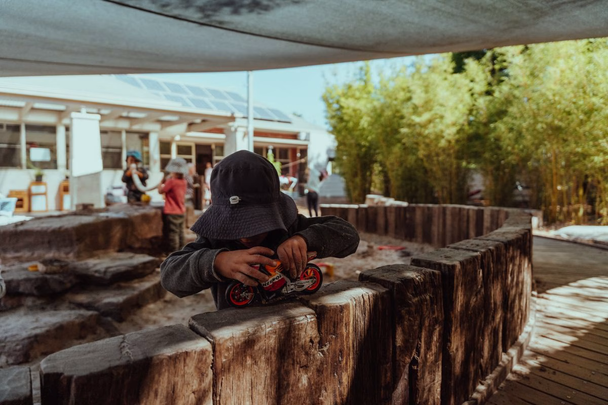 child-playing-outside-with-a-toy-motorbike-on-edge-of-sandpit.avif