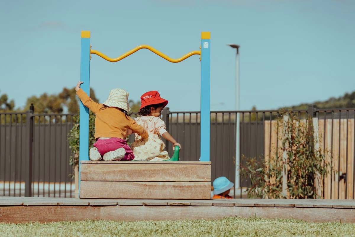 two-childrens-sitting-on-playground-equipment-with-backs-to-us.avif